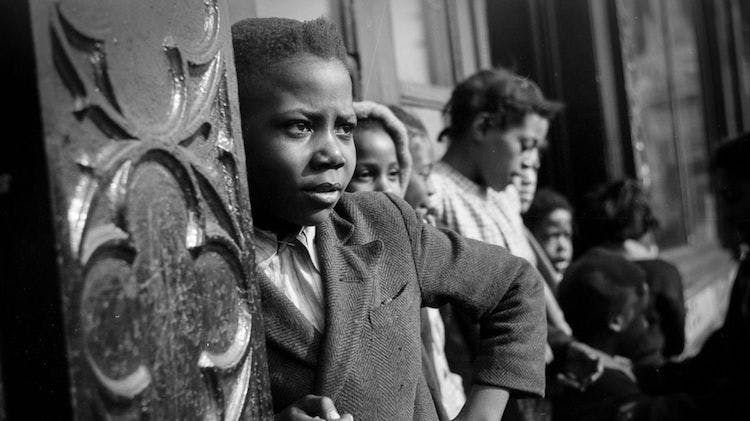A black and white photo taken in 1941 showing a handful of Black children standing in line outside a building. The closest child fills much of the frame, and he looks off into the distance.