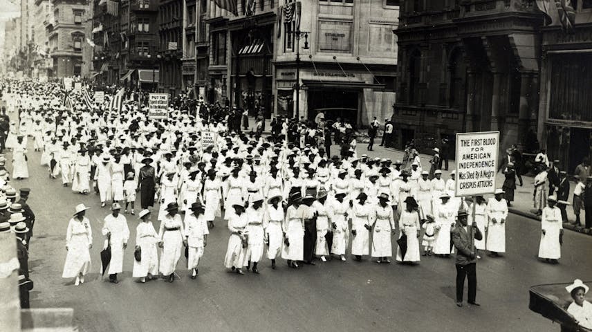 A black-and-white historical photograph showing hundreds of African American women dressed in white marching down a crowded city street in a peaceful protest parade. Some marchers carry signs, including one that reads "The First Blood for American Independence Was Shed By a Negro — Crispus Attucks." American flags hang from the buildings lining the street, and bystanders watch from the sidewalks. This is the Silent Parade of 1917, one of the first major civil rights demonstrations in U.S. history, organized in New York City to protest racial violence and lynching against Black Americans.