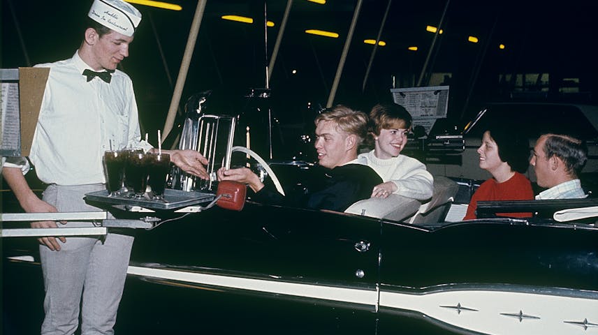 A carhop waiter in a white uniform and paper hat carries a tray of drinks to four smiling teenagers sitting in a black convertible car at a drive-in restaurant at night.