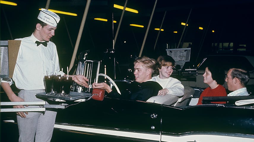 A carhop waiter in a white uniform and paper hat carries a tray of drinks to four smiling teenagers sitting in a black convertible car at a drive-in restaurant at night.