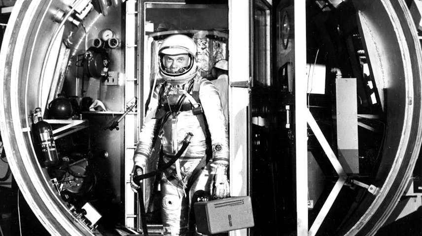 A black and white photo of astronaut John Glenn in a silver pressure suit and helmet, stepping out of a large circular hatch of his Mercury spacecraft in a space simulation chamber. The astronaut is carrying a small rectangular case and is surrounded by scientific equipment and machinery.
