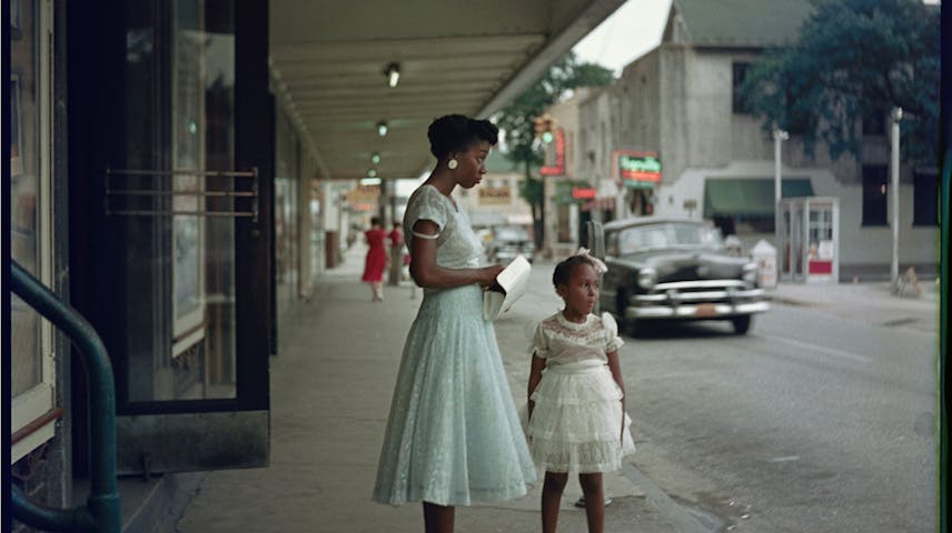 A Black woman in an elegant light blue dress and a young Black girl in a white dress stand on a sidewalk outside a store in Alabama in 1956. Above them, a glowing neon sign reads "Colored Entrance" — a reminder that during racial segregation, Black Americans were forced to use separate doors, waiting areas, and facilities from white people. Both figures are dressed beautifully, yet they face this sign of inequality. The photo was taken by Gordon Parks, a famous Black photographer who documented the injustice of segregation in the United States.