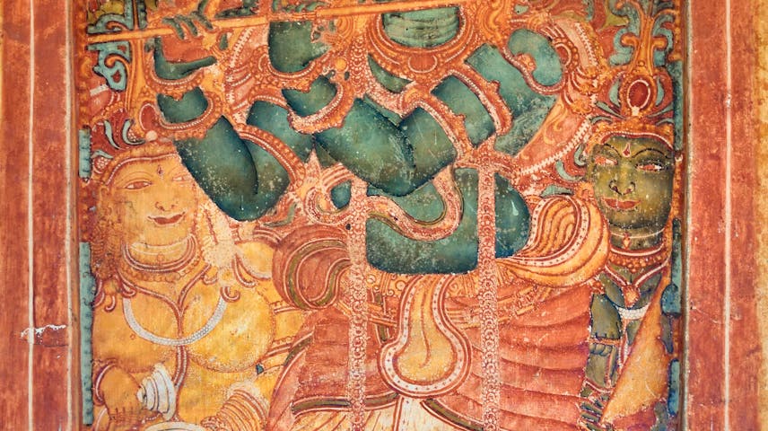 A colorful ancient wall painting from India showing the Hindu god Krishna playing a flute. Krishna is painted with blue-green skin, wearing a tall decorative crown and traditional clothing with lots of jewelry. Two smaller figures stand on either side of him. The painting uses bold shades of red, orange, gold and teal and shows signs of age with some areas faded or chipped. This style of artwork is known as Kerala mural painting and is found on the walls of temples in southern India.