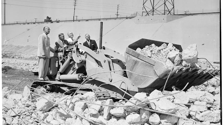 A black-and-white photo from 1957 showing four men in business suits standing on a large bulldozer at the Santa Monica Freeway groundbreaking. The machine's bucket is filled with broken chunks of concrete and rubble.