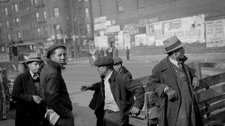 A black and white photo taken in 1941 showing five Black men on the edge of a wide city street, some leaning on a wheeled wooden cart. The men are dressed in work clothes and hats and two of them have glanced at the camera.