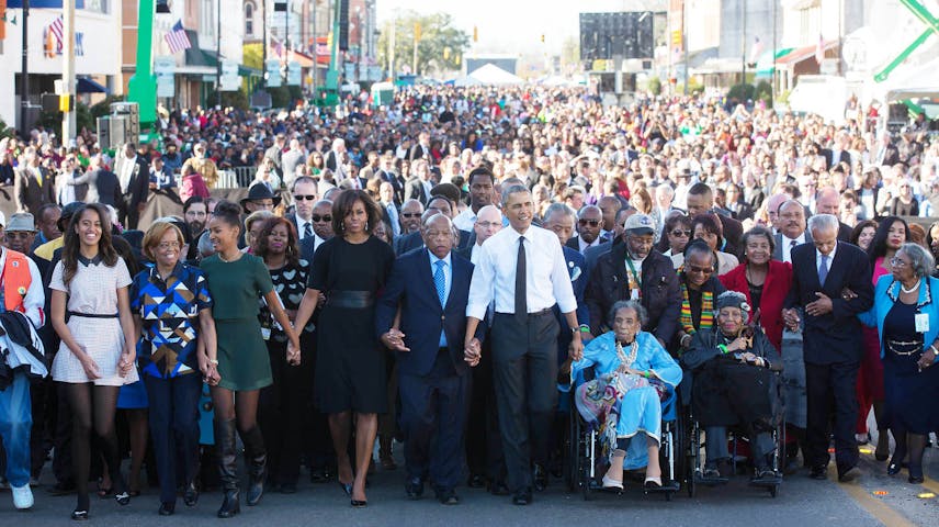 A massive crowd of people march together down a city street, filling it from side to side as far as the eye can see. In the front row, people of different ages hold hands, including an elderly woman in a wheelchair. The group walks in a spirit of unity and purpose, reminiscent of the historic Civil Rights marches of the 1960s.