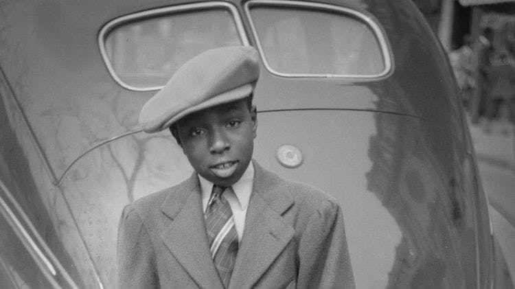 A black and white portrait photo taken in 1941 of a Black boy wearing a suit, tie and a large newsboy-style hat. He is leaning on the back of a car