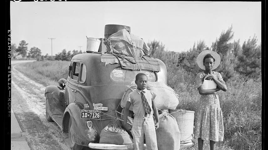 A black and white photo of two Black children standing behind a car loaded on top with their belongings.