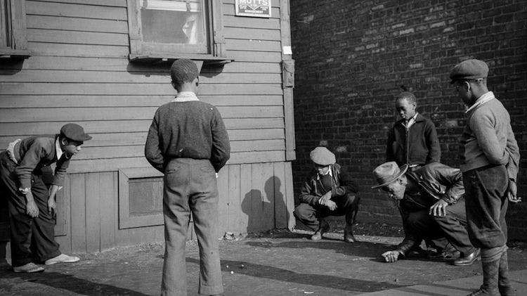 A black and white photo taken in 1941 of five Black boys and one adult man. They stand in a circle and the man is shooting marbles while the boys watch intently.