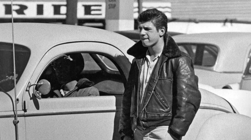 A black and white photo of a young man leaning against a 1950s car in a parking lot. He is wearing a leather jacket and light-colored pants and is holding a cigarette. His hair is styled in a slicked-back look typical of the 1950s. Other cars and a sign can be seen in the background.