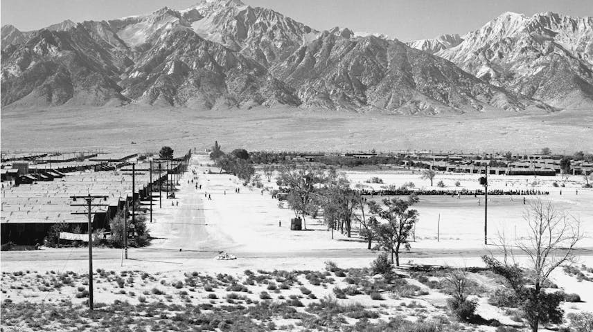 A black-and-white photograph taken from a guard tower looking down at the Manzanar War Relocation Center, a Japanese American incarceration camp in California during World War II. Rows of long, low wooden barracks stretch across a flat, dusty desert landscape. A wide dirt road runs through the center of the camp, with telephone poles lining each side and sparse desert shrubs and bare trees scattered throughout. In the distance, a large crowd of people can be seen gathered near the edge of the camp. Towering snow-capped mountains of the Sierra Nevada rise dramatically against a clear sky in the background. The photo was taken by the famous photographer Ansel Adams in 1943.