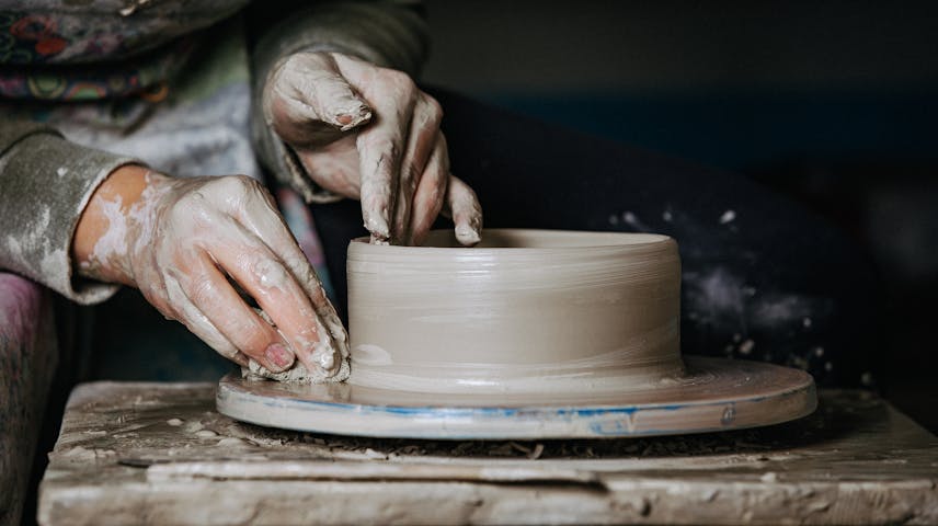 Image of two hands shaping a bowl on a pottery wheel; one hand is positioned inside the bowl, and the other is at its base, holding a small sponge.