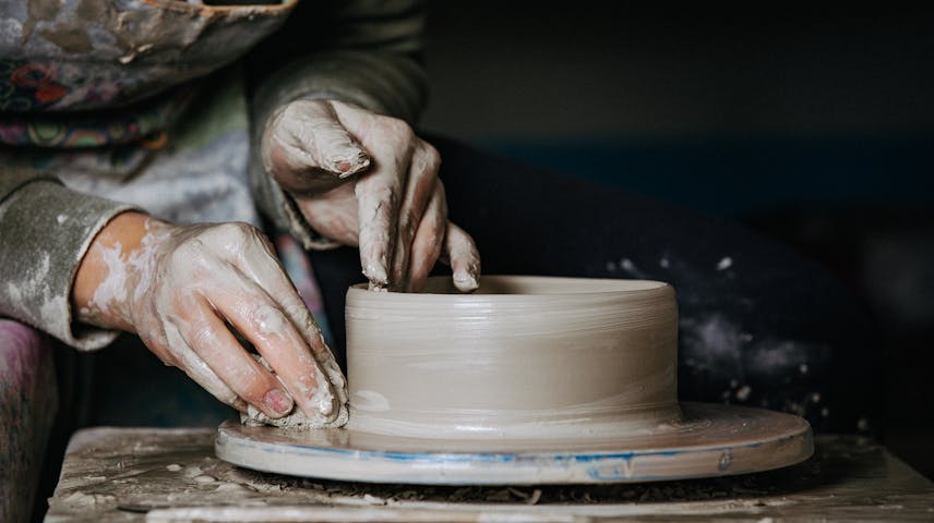 Image of two hands shaping a bowl on a pottery wheel. One hand is positioned inside the bowl, the other is at its base holding a small sponge.