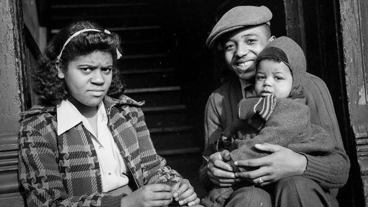 A black and white photo taken in 1941 showing what appears to be a Black couple. They are sitting on the front stoop of a building and he is holding a toddler. The man has a big smile but the woman’s expression is more guarded.