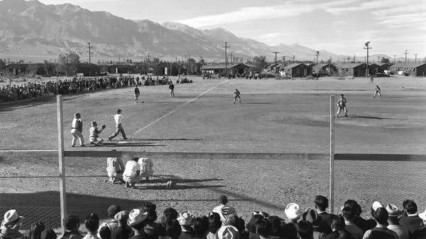 A black-and-white photograph of a baseball game being played on a dirt field, with a large crowd of spectators watching from behind a fence in the foreground. Players are visible at home plate and spread across the field. In the background are rows of small, plain buildings and tall mountains under a wide open sky.