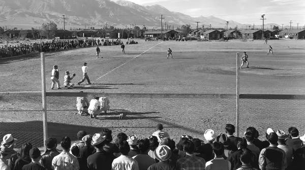 A black-and-white photograph of a baseball game being played on a dirt field, with a large crowd of spectators watching from behind a fence in the foreground. Players are visible at home plate and spread across the field. In the background are rows of small, plain buildings and tall mountains under a wide open sky.