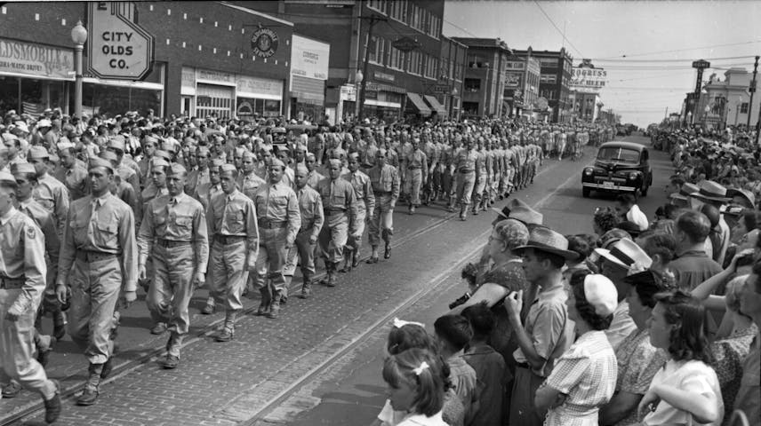 A black and white historical photo showing a large group of soldiers in uniform marching in rows down a brick-paved city street while crowds of civilians line the sidewalk to watch. Storefronts and signs, including one for “City Olds Co.,” are visible in the background. The clothing and cars show this parade took place in the 1940s.