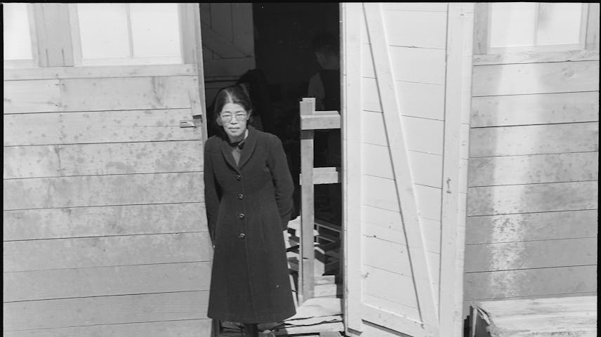 A black-and-white photograph of a Japanese American woman standing in the doorway of a small wooden barrack-style building. She is wearing a long dark coat, boots, and glasses, and is looking at the camera. The building appears to be hastily constructed, with rough wooden boards and a dirt ground visible. This photo was taken in San Bruno, California, at the Tanforan Assembly Center, where Japanese Americans were forced to live temporarily during World War II after the U.S. government ordered their removal from the West Coast following the attack on Pearl Harbor.