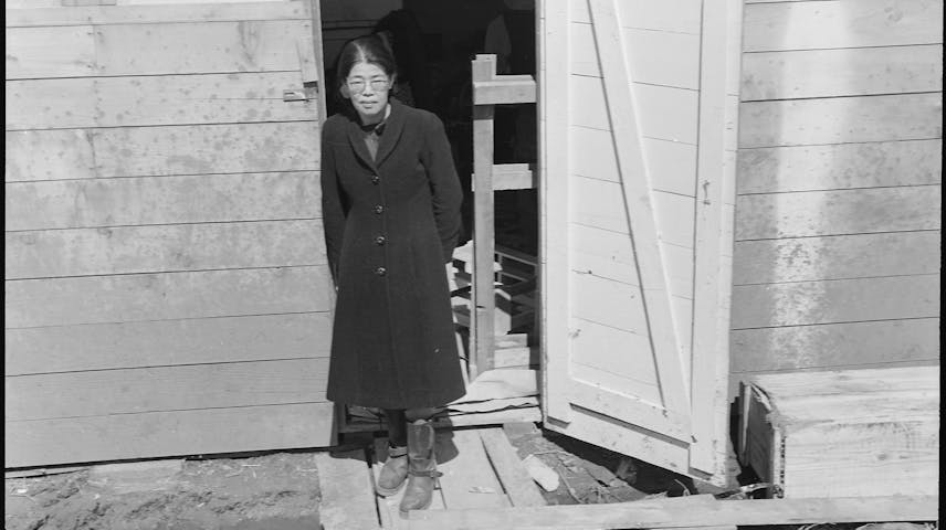 A black-and-white photograph of a Japanese American woman standing in the doorway of a small wooden barrack-style building. She is wearing a long dark coat, boots, and glasses, and is looking at the camera. The building appears to be hastily constructed, with rough wooden boards and dirt ground visible. This photo was taken in San Bruno, California, at the Tanforan Assembly Center, where Japanese Americans were forced to live temporarily during World War II after the U.S. government ordered their removal from the West Coast following the attack on Pearl Harbor.