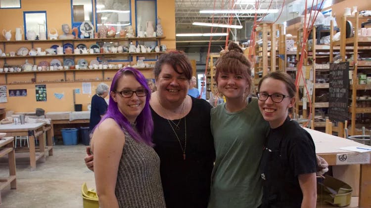 Four smiling people pose together in a pottery studio. Shelves of clay pots, plates, and sculptures fill the walls behind them. One person has bright purple hair. They look happy and proud of the creative workspace around them.