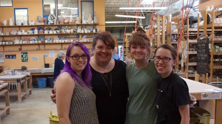 Four smiling people pose together in a pottery studio. Shelves of clay pots, plates, and sculptures fill the walls behind them. One person has bright purple hair. They look happy and proud of the creative workspace around them.