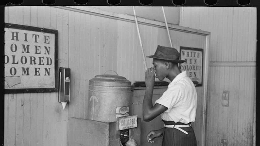 A black man drinking at a metal water cooler with a sign that says “Colored” on it.