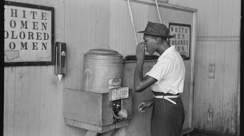 A black man drinking at a metal water cooler with a sign that says “Colored” on it.