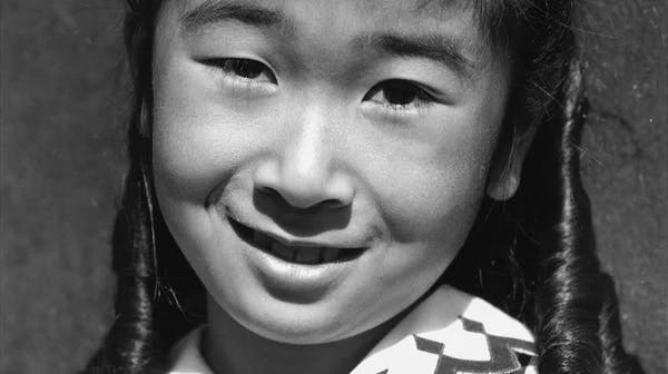 Black-and-white photograph of a young Japanese American girl smiling at the camera. She has dark hair in two pigtails and is wearing a patterned button-up shirt with stripes and geometric designs. The photo was taken by famous photographer Ansel Adams at Manzanar, a Japanese American incarceration camp during World War II.