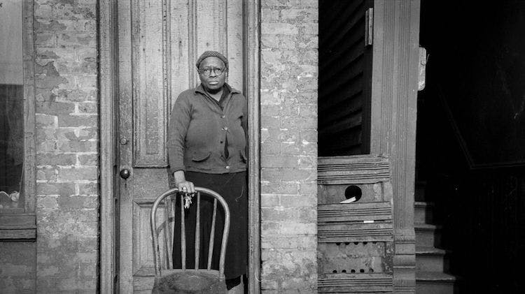 A black and white photo taken in 1941 showing a Black woman standing in the doorway of a brick building. She holds the back of a wooden chair that is in front of her. She is wearing a long skirt, a partially buttoned sweater, glasses and is not smiling.