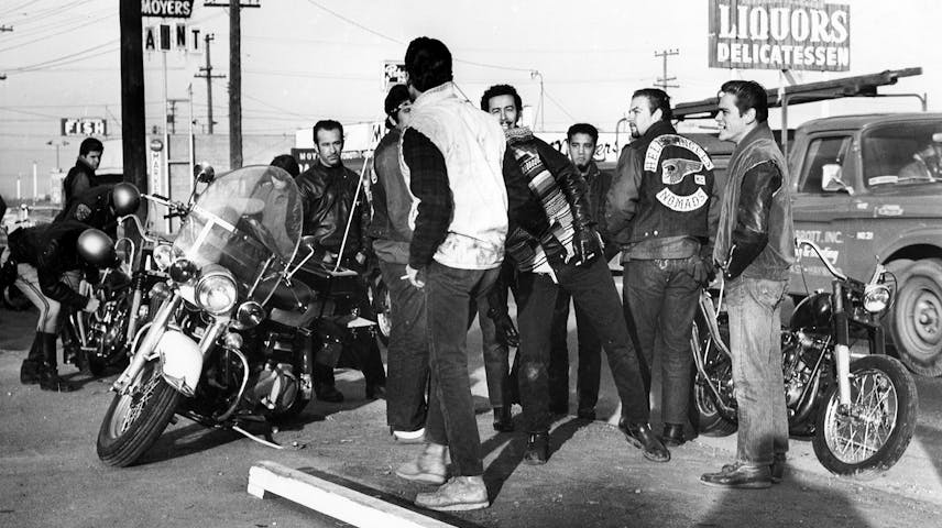 A black and white photograph from the 1960s showing a group of young men who are part of the Hell’s Angels motorcycle club gathered in a parking lot with motorcycles. Several of the men are wearing leather jackets. There is one police officer visible who is examining a motorcycle. In the background, there are street signs, power lines and store signs.