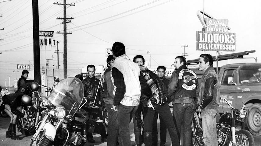 A black and white photograph from the 1960s showing a group of young men who are part of the Hell’s Angels motorcycle club gathered in a parking lot with motorcycles. Several of the men are wearing leather jackets. There is one police officer visible who is examining a motorcycle. In the background, there are street signs, power lines and store signs.