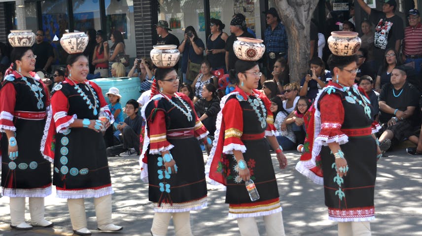 Women in traditional Zuni dress walk down a street during a parade while balancing Zuni pottery on their heads. There are onlookers seated and standing lining the street to watch.