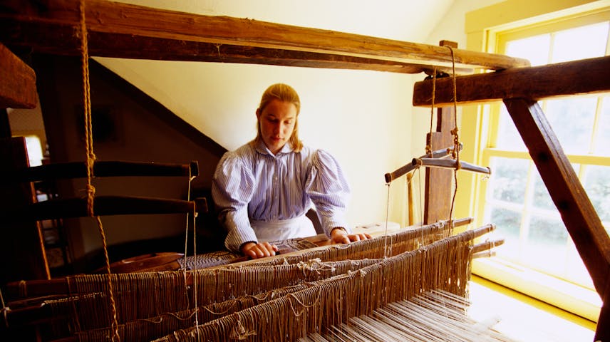 A young woman in old-fashioned clothing operates a large wooden loom, carefully weaving threads into fabric by hand.
