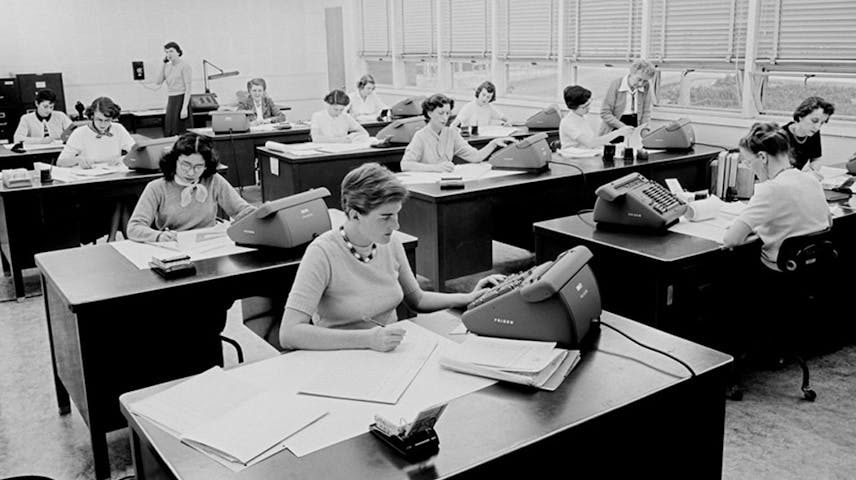 Historical photograph of several women seated at desks with papers, calculators, and office equipment in a workroom.