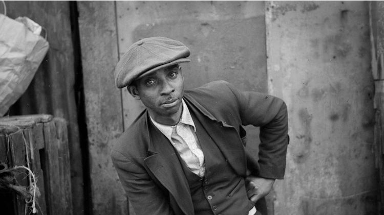 A black and white portrait photo taken in 1941 of a young Black man, seated in front of an exterior wall. He has one hand on his hip and leans on his leg with his other arm. He looks at the camera and is wearing a suit, vest and large newsboy-style hat.