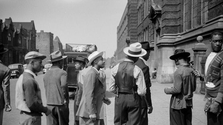 A black and white photo taken in 1941 showing several smartly dressed young black men, none looking right at the camera, standing on a wide city sidewalk with brick buildings and vehicles in the background