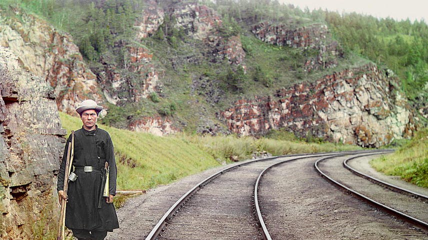 A man with a shovel stands next to train tracks in a mountainous region.
